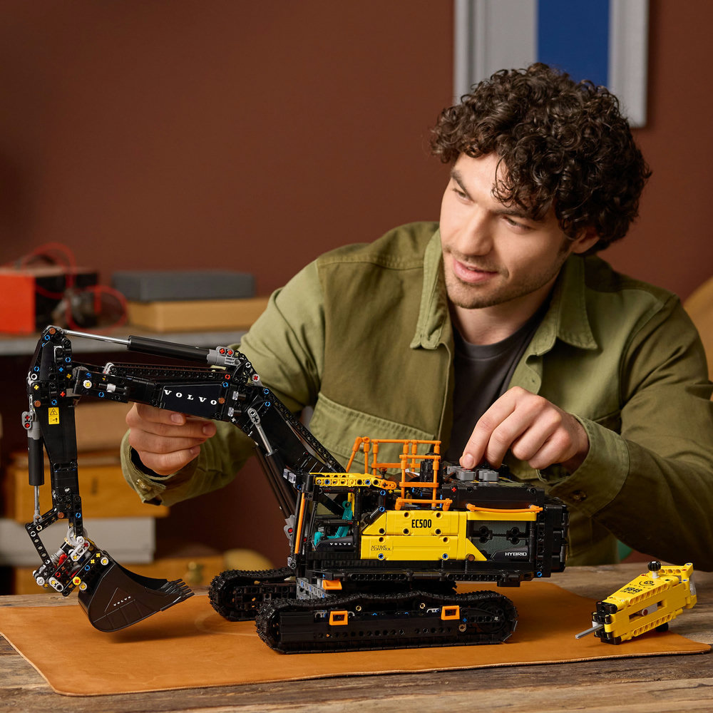 Man playing with a LEGO Technic: Volvo EC500 Hybrid Excavator on a wooden table