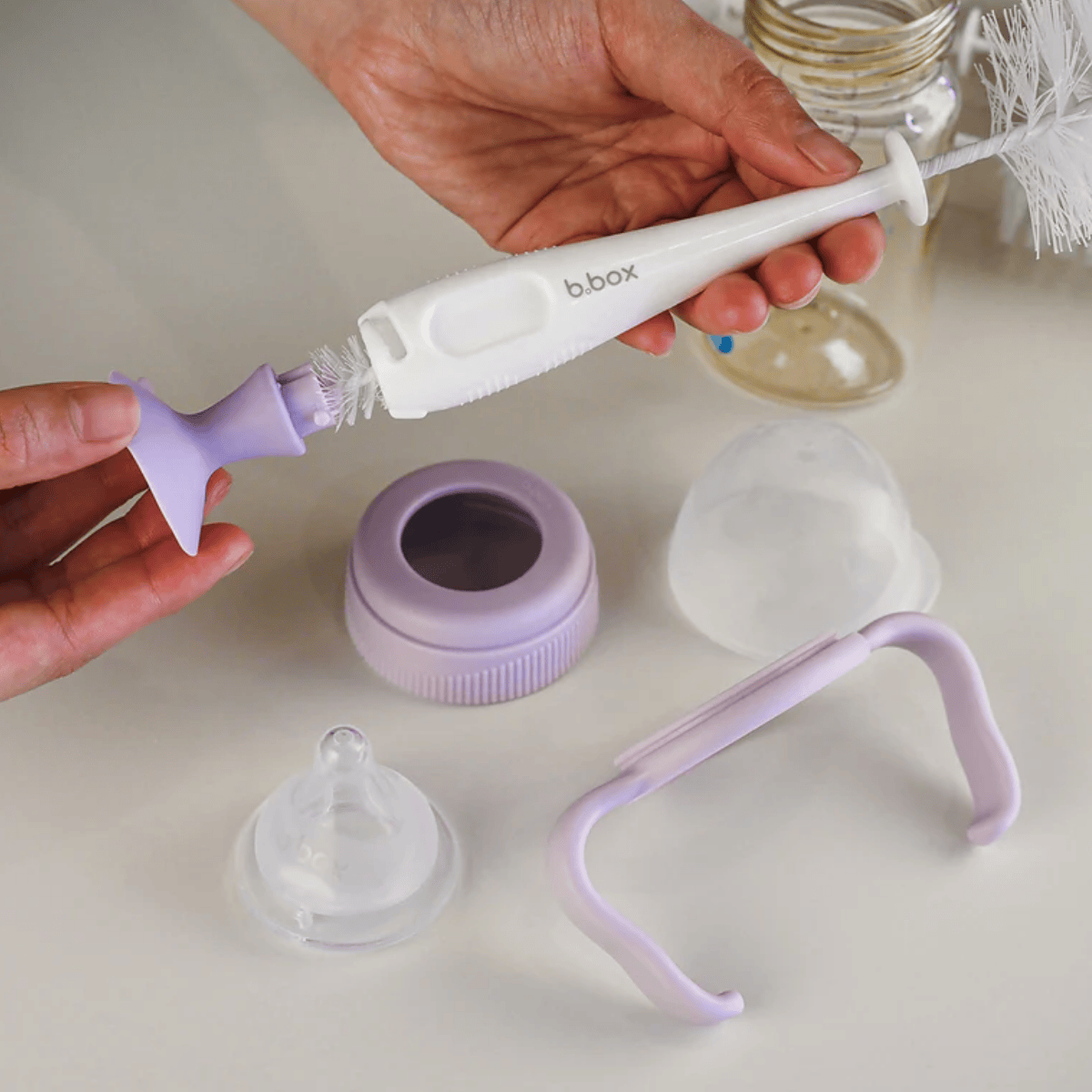Person using a b.box cleaning brush on a baby bottle with various components on a light surface.