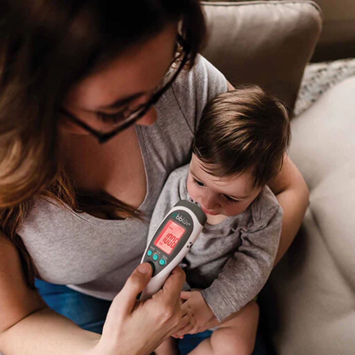 Woman holding a child and using a digital thermometer on a couch