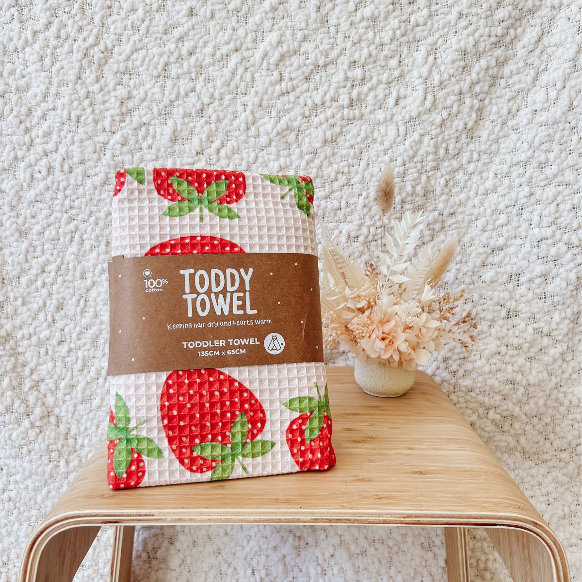Waffle-textured toddler towel with strawberry design on a wooden stool against a textured white wall.