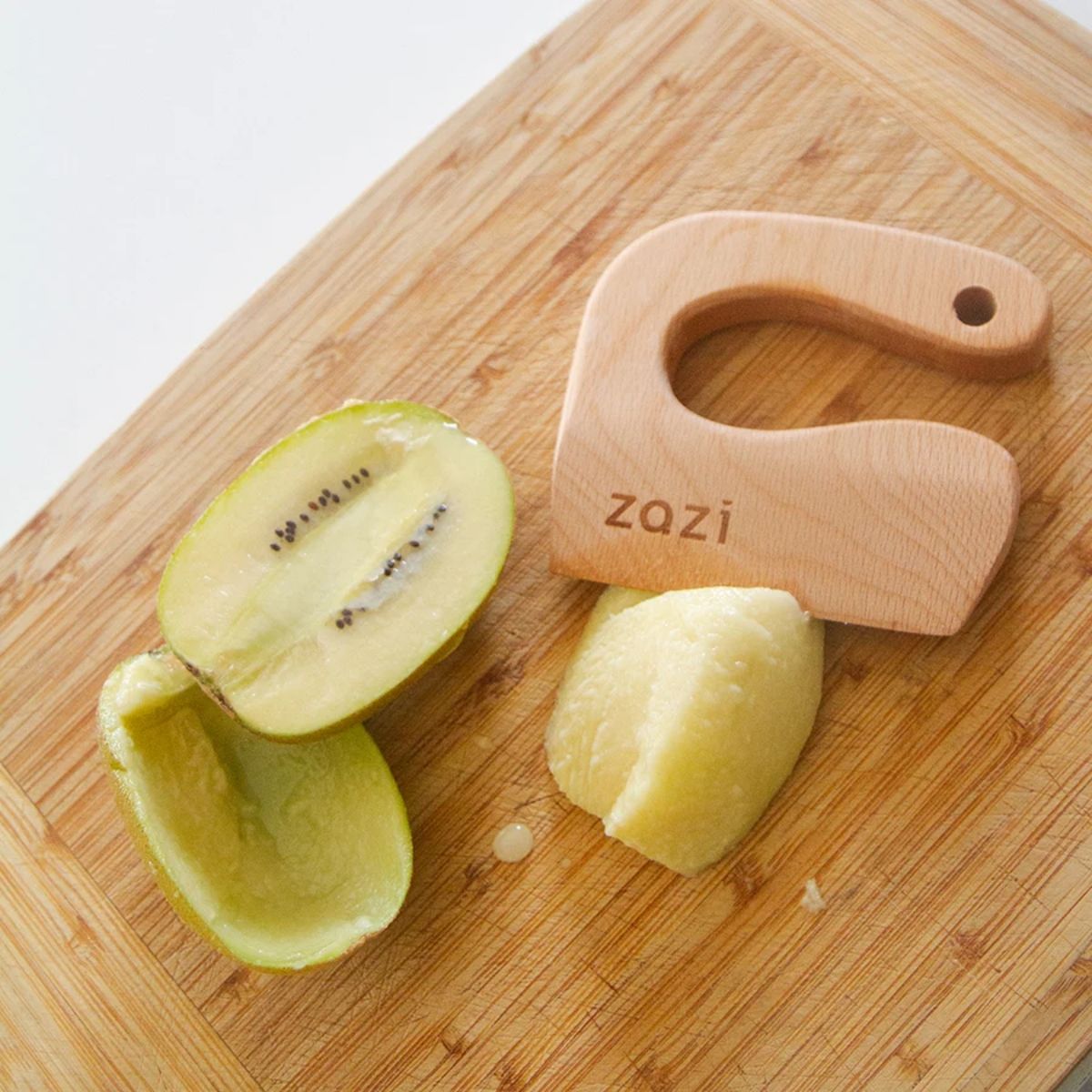 Wooden knife with 'zazi' branding on a wooden cutting board with a halved kiwi and a piece of peeled apple.