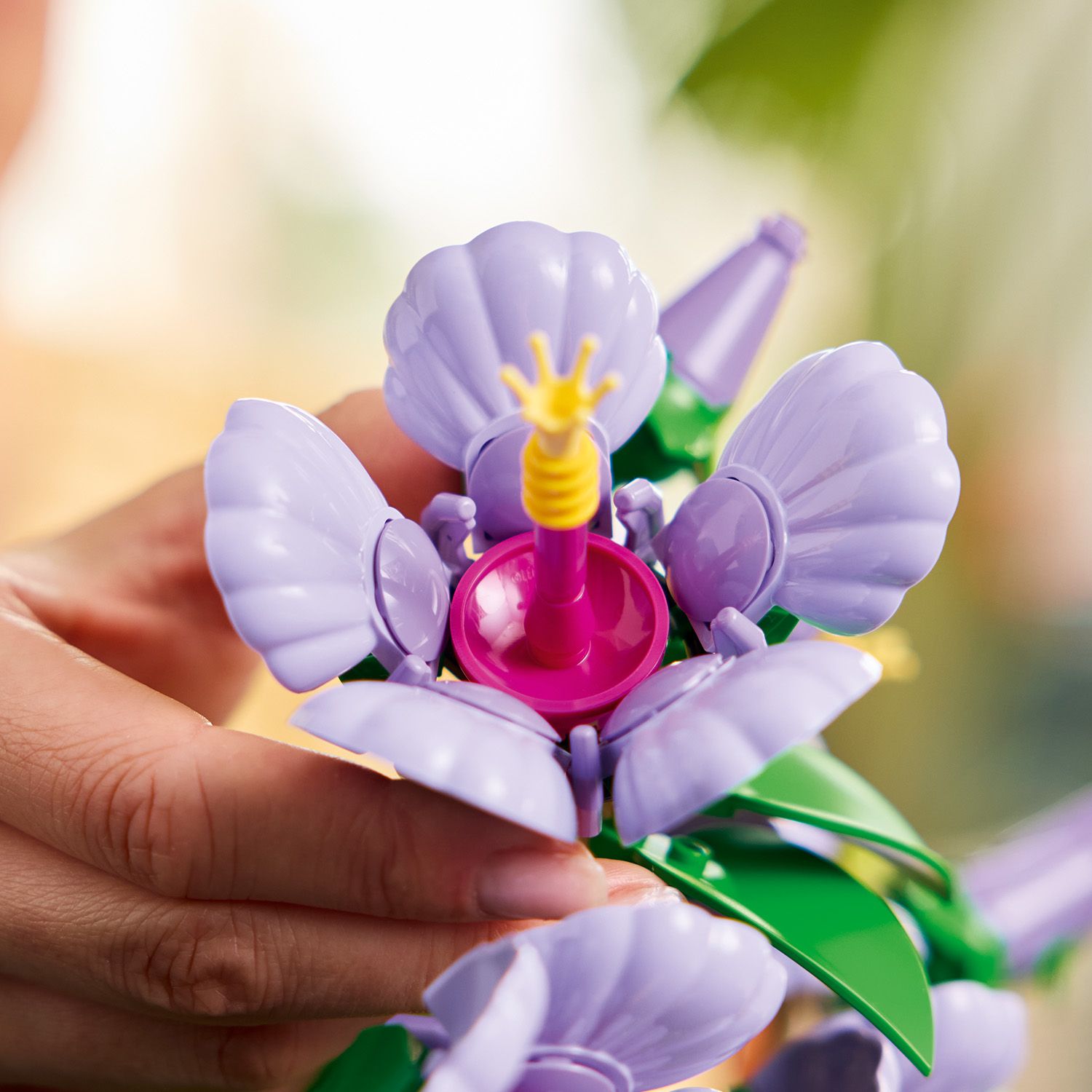 LEGO Toy figure of a flower with purple petals and a pink center held by a hand against a blurred background.