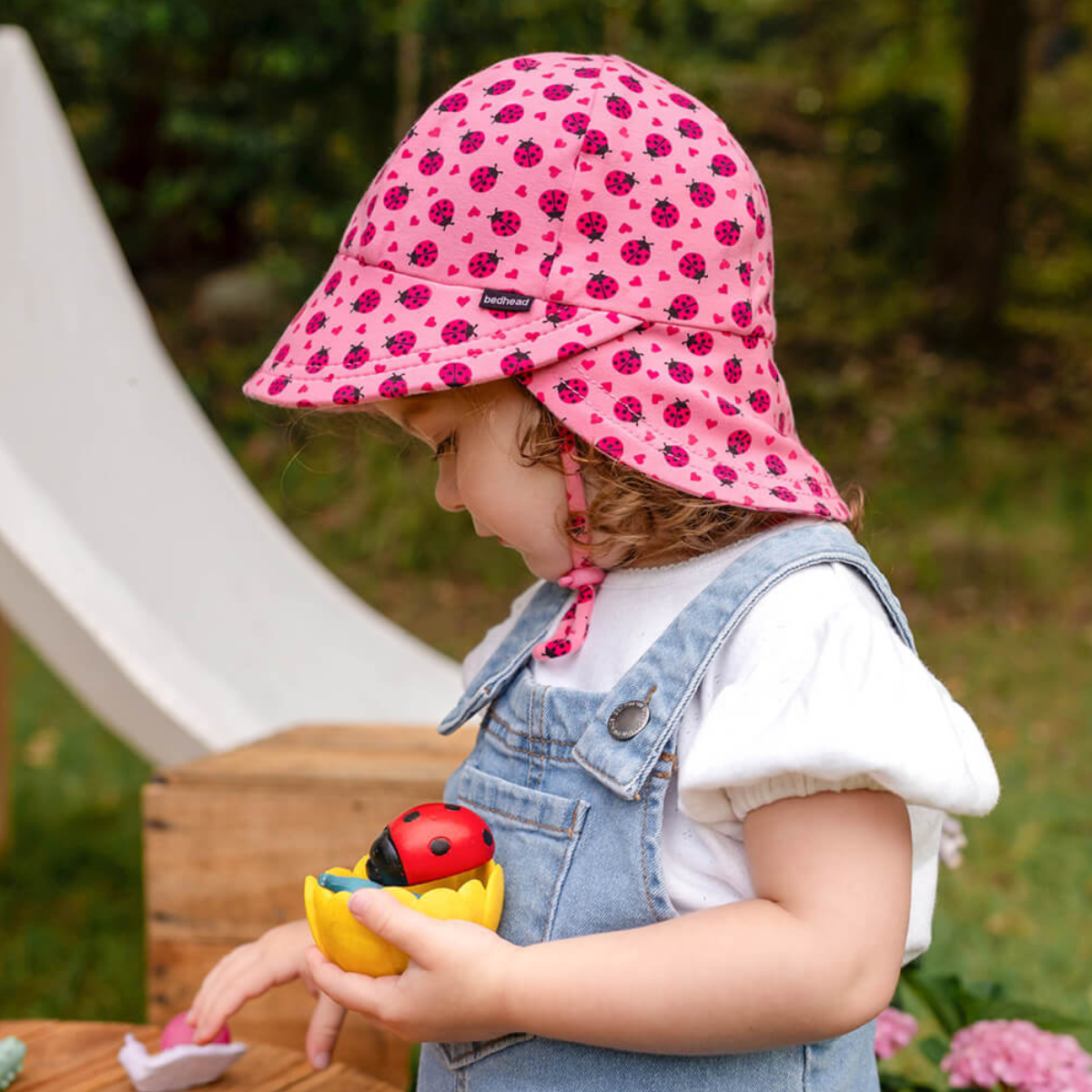 Child wearing a pink sun hat with ladybug pattern, holding a toy duck with a ladybug on it, outdoors.