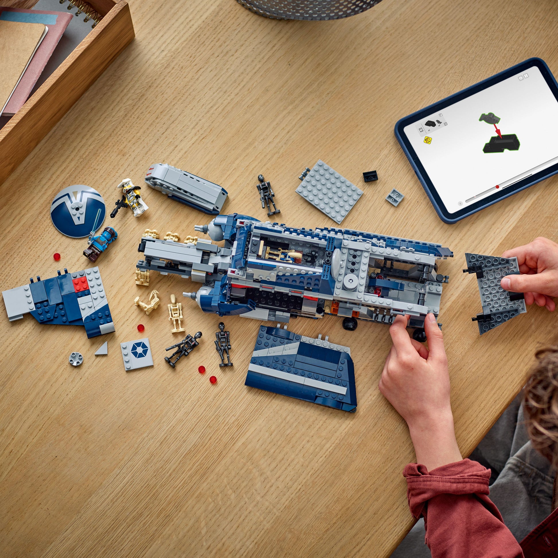 Person assembling a large blue and gray Lego model on a wooden table with a tablet displaying building instructions.