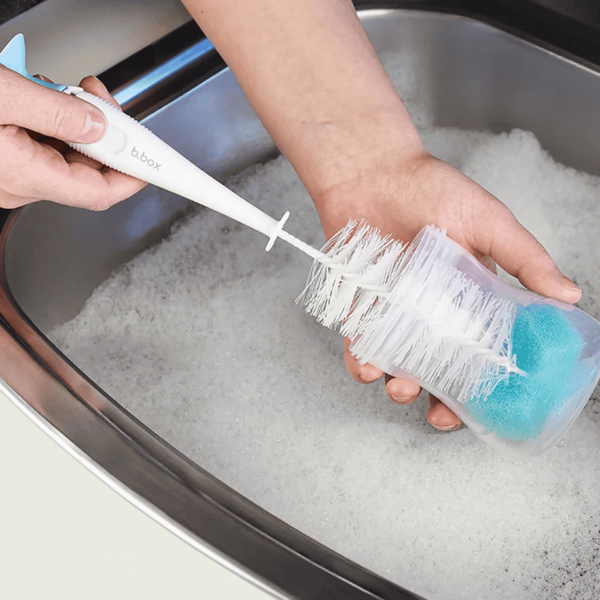 Person cleaning a bottle brush with a blue sponge in a sink filled with soapy water.
