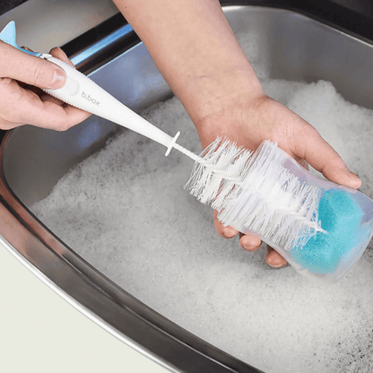 Person cleaning a bottle brush with a blue sponge in a sink filled with soapy water.