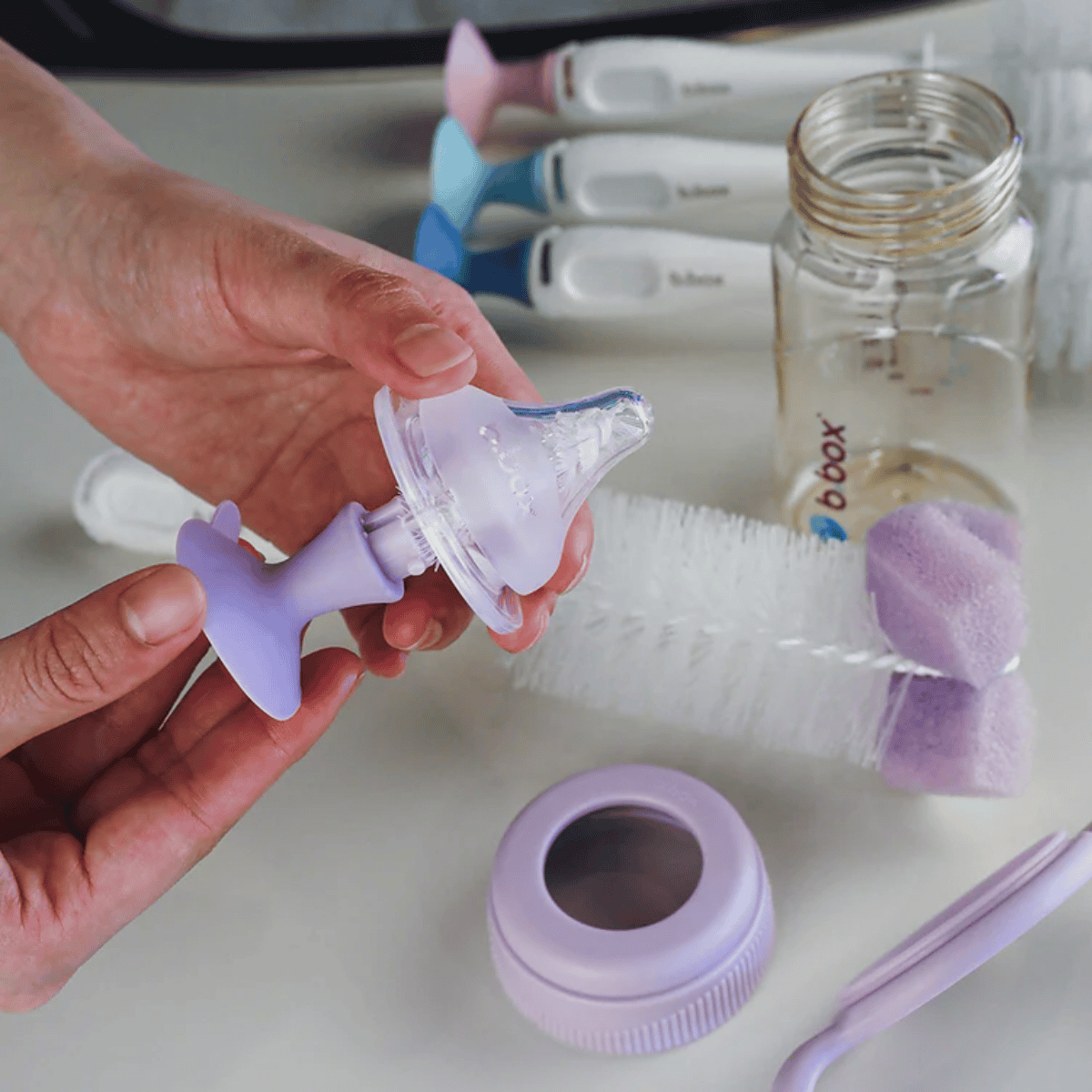 Hand holding a baby bottle nipple with cleaning brush and bottle in the background