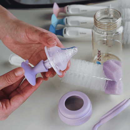 Hand holding a baby bottle nipple with cleaning brush and bottle in the background