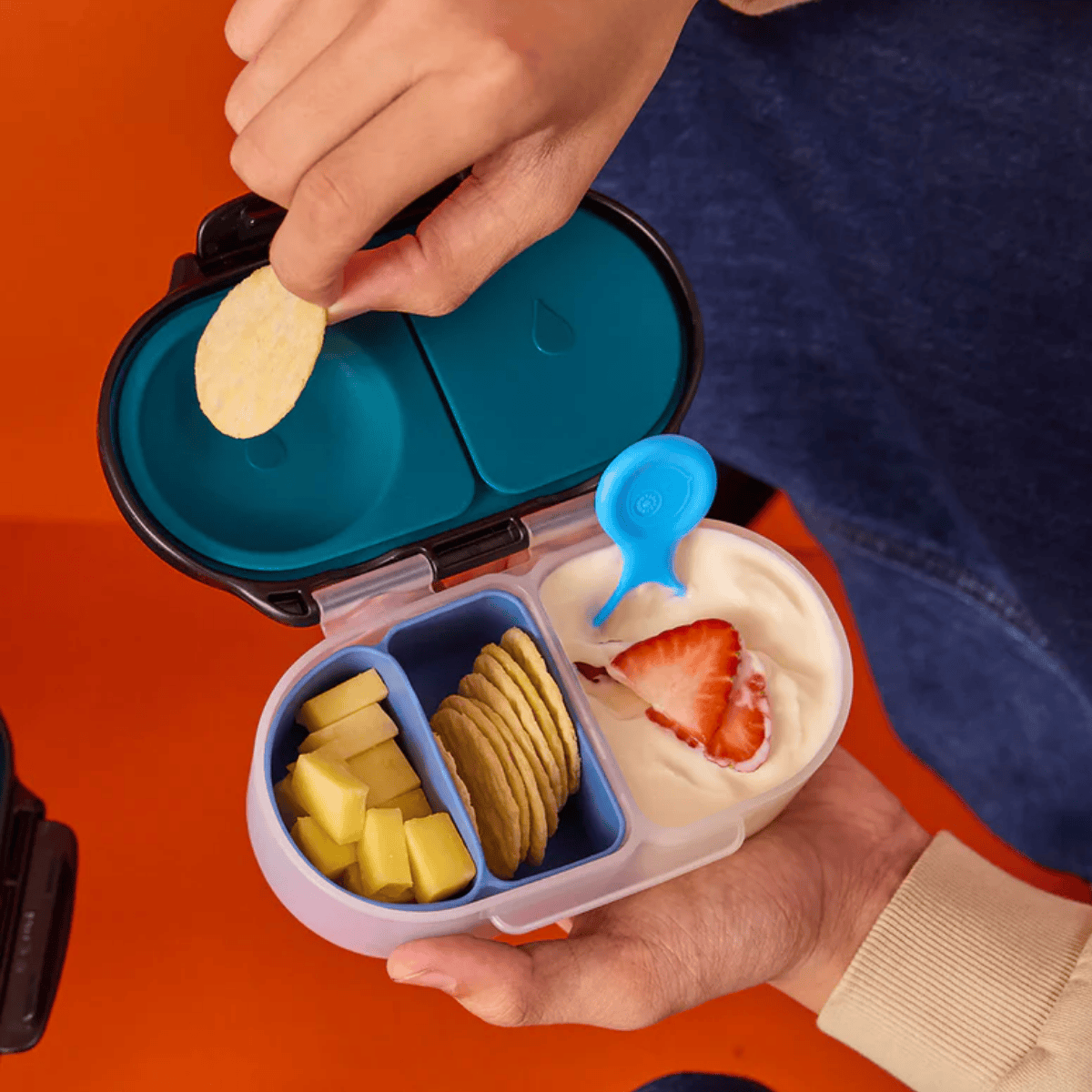Person opening a small, openable container with compartments filled with snacks on an orange background.