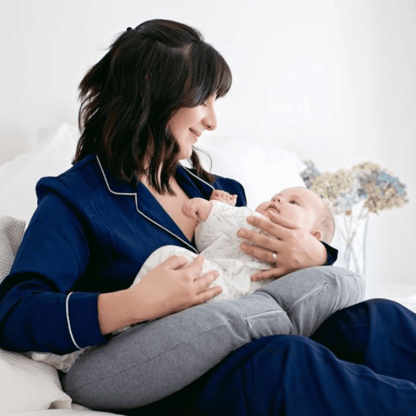 Woman in blue pajamas holding a baby on a bed surrounded by a breastfeeding cushion