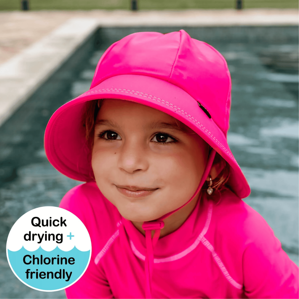 A young child wearing a bright pink sun hat and matching pink swim top, photographed outdoors near a pool. A circular badge in the lower left reads 'Quick drying + Chlorine friendly'.