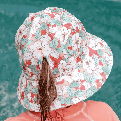 A close-up back view of a child wearing a wide-brimmed sun hat with a white, coral, and mint floral print, paired with a coral long-sleeve top, set against a teal water background.