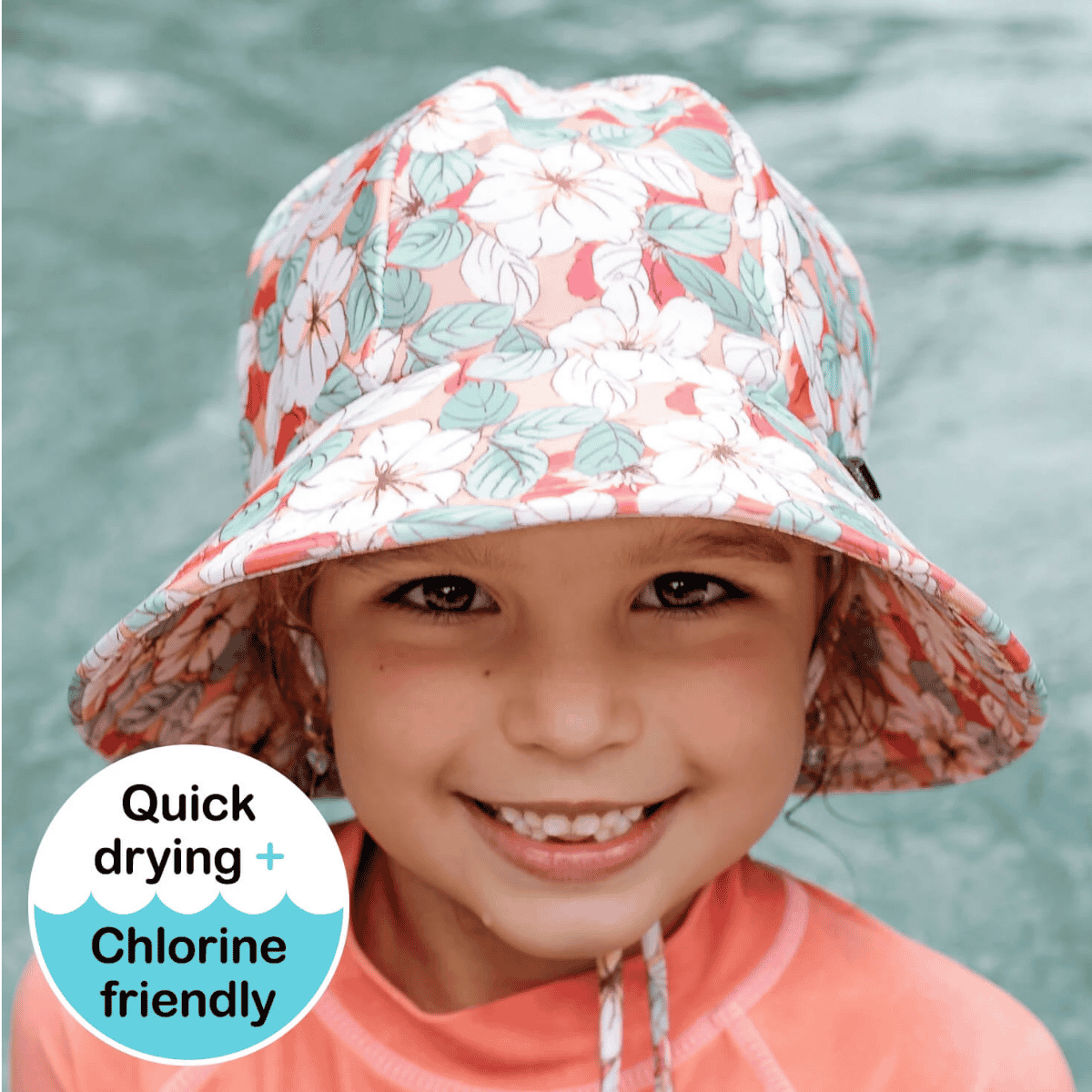 A smiling child wearing a floral bucket sun hat with a coral, white, and light green pattern, photographed outdoors against a blurred water background. A circular badge in the lower left reads 'Quick drying + Chlorine friendly'.
