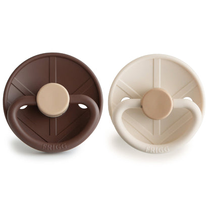 Two baby pacifiers, one brown and one beige, on a white background with 'FRIGG' branding, which is a Swedish Brand.