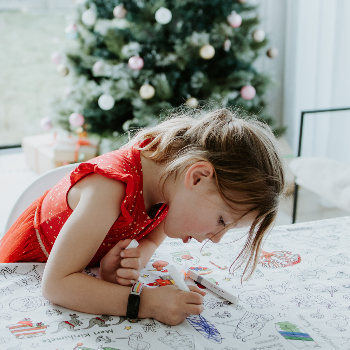 Child in a red dress coloring on a washable table cloth with a Christmas tree in the background