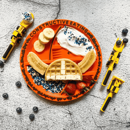 An overhead flat-lay of a children's waffle-themed plate on a textured gray surface, surrounded by toy construction tools. The orange plate has compartments with waffles, banana slices, strawberries, whipped cream, blue sprinkles, and the words 'CONSTRUCTIVE EATING'.