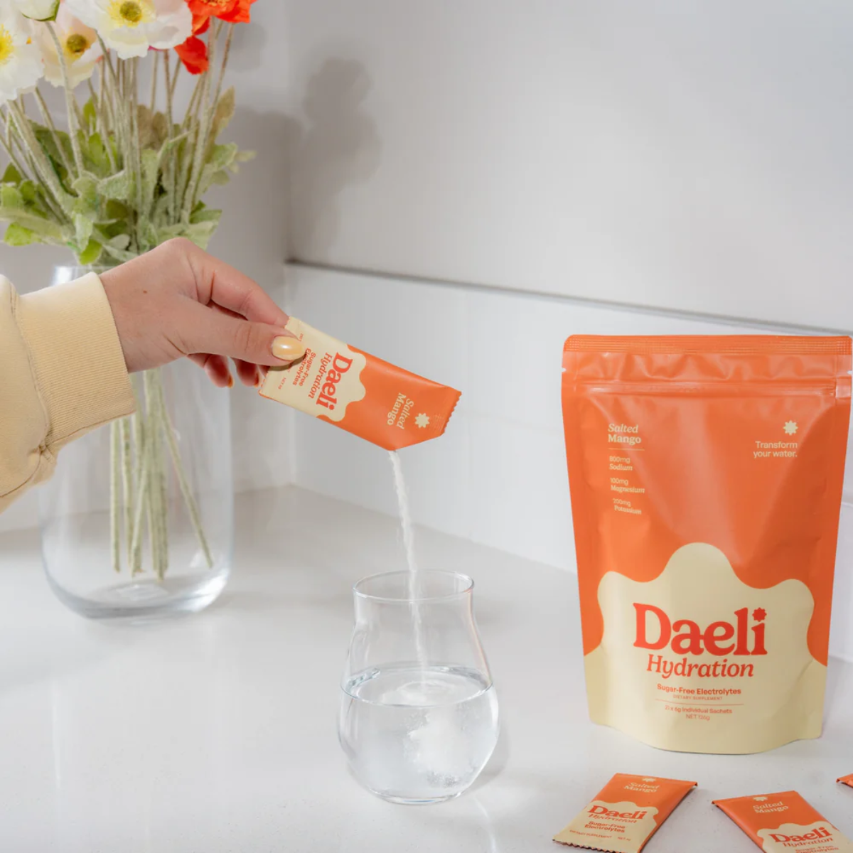 Person holding a Daeli hydration packet with a large package and glass on a white surface.