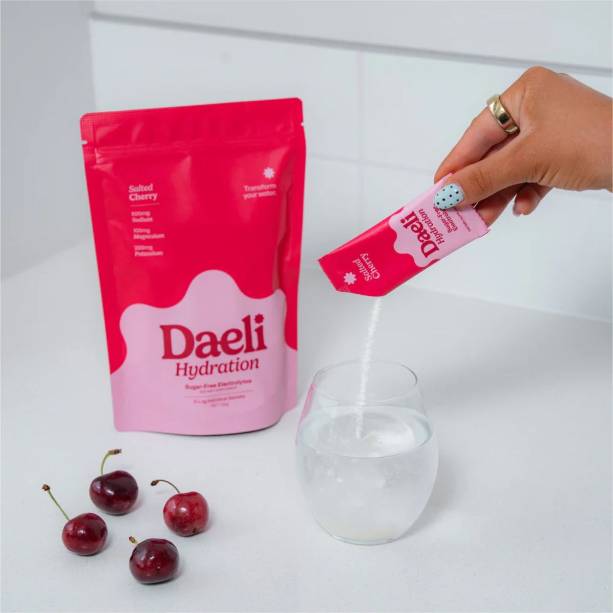 Daeli Hydration packaging with a hand pouring a pink packet into a glass of water, surrounded by cherries on a white background.