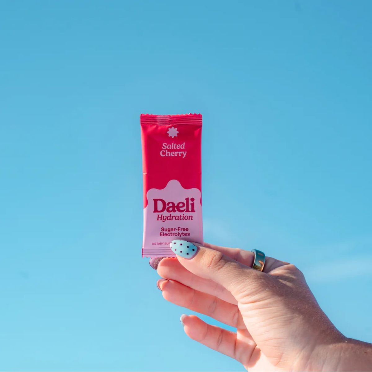 Hand holding a pink Daeli hydration package against a blue sky