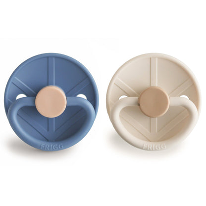 Two pacifiers, one blue and one beige, on a white background with 'FRIGG' branding.