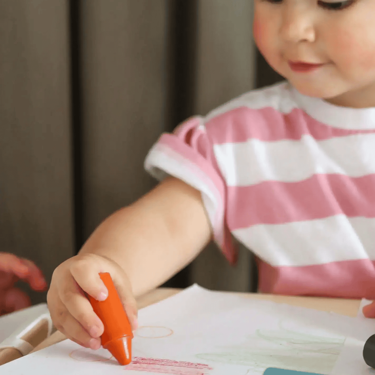 Child holding an orange crayon over a piece of paper with drawings.