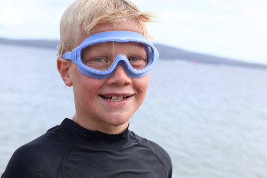 Child wearing blue goggles and a black wetsuit with a blurred water background