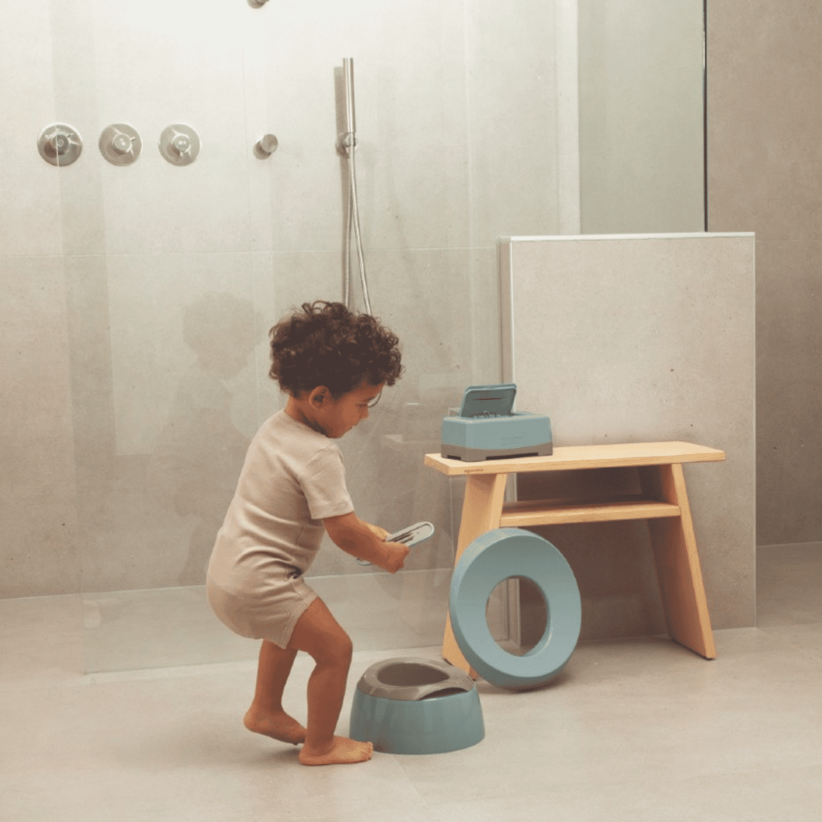 A toddler stands in a bathroom next to a wooden stool, holding a small light blue potty training seat and ring, with a matching seat on the floor.