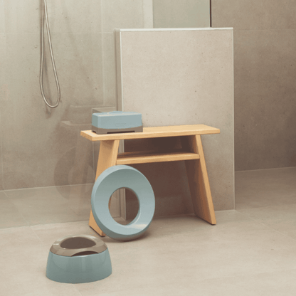 A minimalist bathroom scene with a light wood stool, a light blue ring-shaped toilet lid, and a matching light blue toilet bowl on a beige tiled floor.