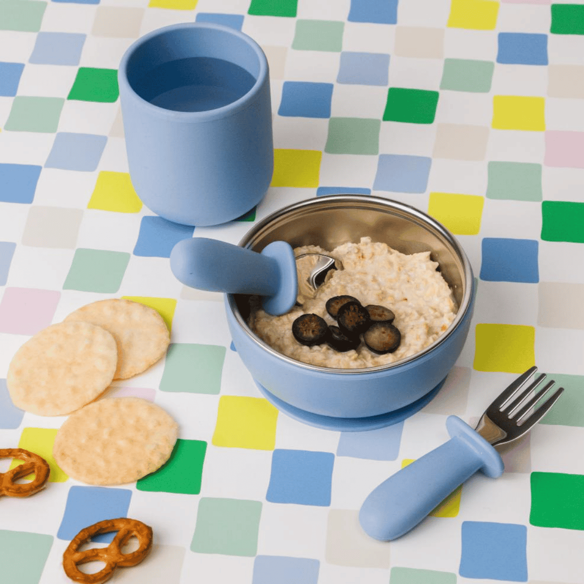 Children's blue bowl, cup, and fork set on a checkered tablecloth with food.