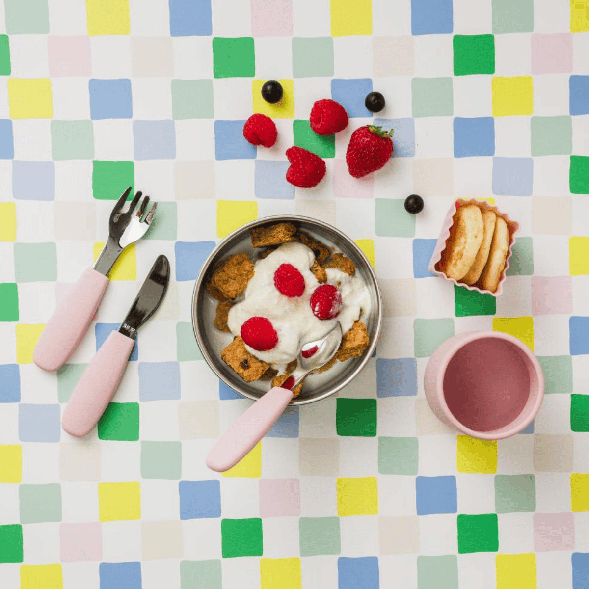 Breakfast setting with a bowl of cereal, yogurt, and berries on a colorful checkered tablecloth.