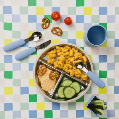 A colorful children's divided lunch tray filled with macaroni, cucumber slices, crackers, and pretzels, styled on a pastel checkered tablecloth with blue-handled utensils and a blue cup.