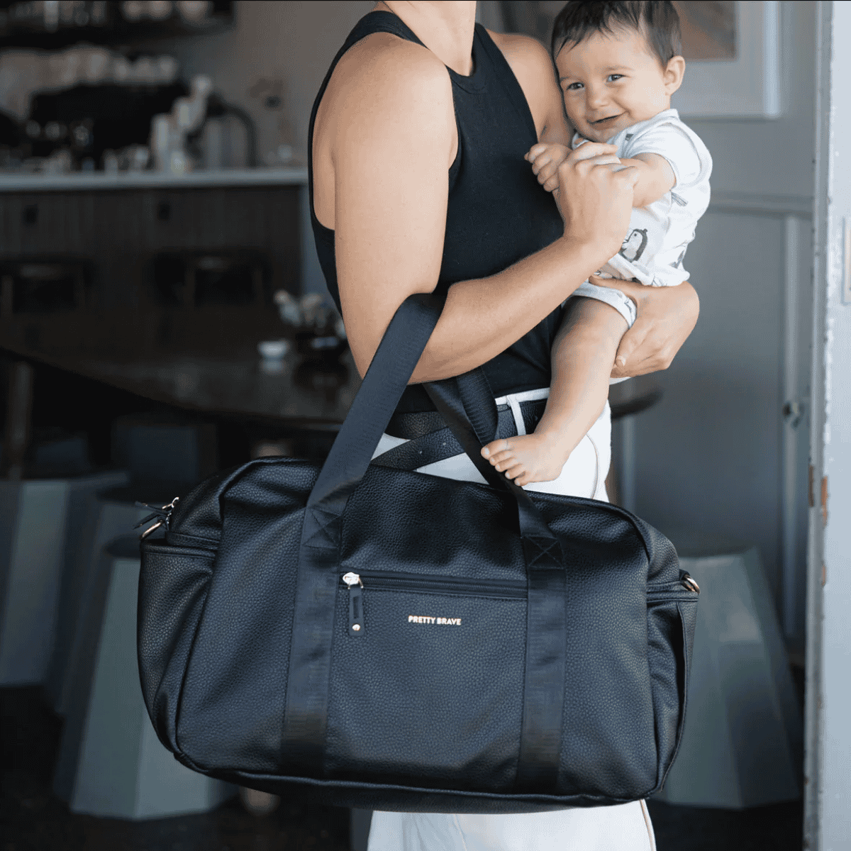 Person holding a baby and a black duffel bag with 'Pretty Brave' branding in an indoor setting.