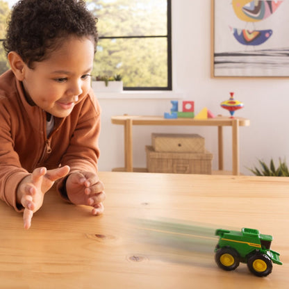 Child playing with a John Deere toy tractor on a wooden table in a room with toys and a window in the background.